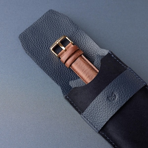 May include: A brown leather watch band with a gold buckle is partially visible inside a dark blue leather watch case. The case is open, revealing the watch band. The case has a textured finish and a strap closure.