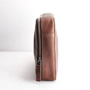 May include: A brown leather zippered pouch with a silver zipper. The pouch is closed and standing upright on a white surface.