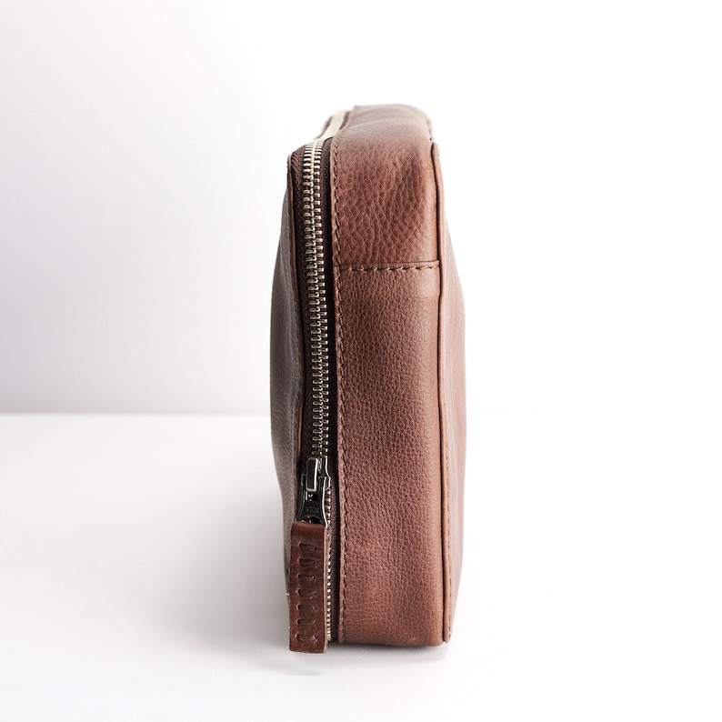 May include: A brown leather zippered pouch with a silver zipper. The pouch is closed and standing upright on a white surface.