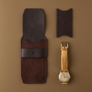 May include: A watch with a light brown leather band and gold-colored case next to a brown leather watch case and a small brown leather sleeve. The watch case is open, revealing a brown interior.