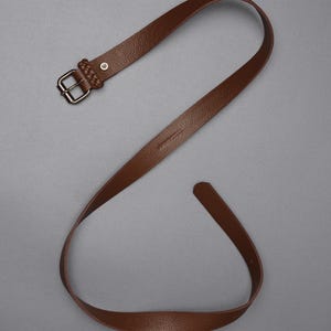 May include: A brown leather belt with a textured surface. The belt features a silver-colored buckle and a braided leather loop near the buckle. The belt is displayed against a gray background.