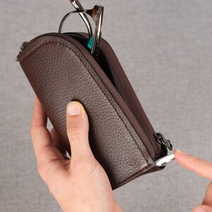 Puede incluir: Un estuche de cuero marr&oacute;n con cremallera para gafas. El estuche tiene un peque&ntilde;o logotipo en la parte delantera y un tirador de cremallera con un anillo plateado.