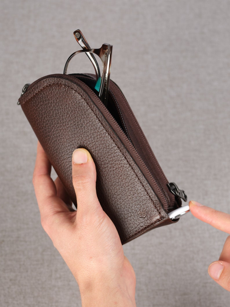 Puede incluir: Un estuche de cuero marr&oacute;n con cremallera para gafas. El estuche tiene un peque&ntilde;o logotipo en la parte delantera y un tirador de cremallera con un anillo plateado.