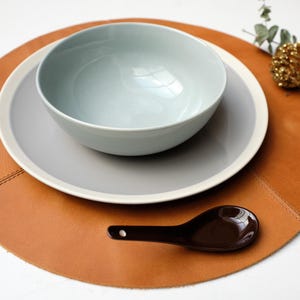Puede incluir: Una mesa con un cuenco azul claro, un plato gris con borde blanco y una cuchara marrón sobre un salvamanteles redondo de cuero marrón. Ramas decorativas y una piña dorada en el fondo.