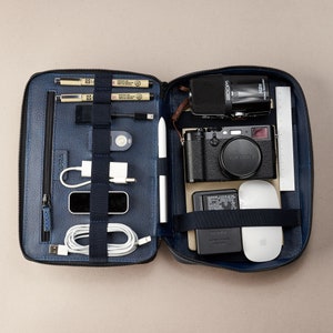 May include: Navy blue leather organiser case, open to reveal tech accessories. Contains a black camera, Zoom H2n recorder, Apple mouse, pens, cables, and a stylus. The case is zipped and has multiple compartments for organisation.