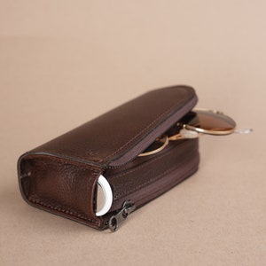 Puede incluir: Un estuche de cuero marr&oacute;n con cremallera para gafas de sol. El estuche est&aacute; abierto y se puede ver parcialmente un par de gafas de sol en el interior.