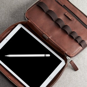 May include: A brown leather zippered case with a white tablet and a white stylus inside. The case has multiple slots for pens or pencils. The case has the word "CARRA" printed on the front.