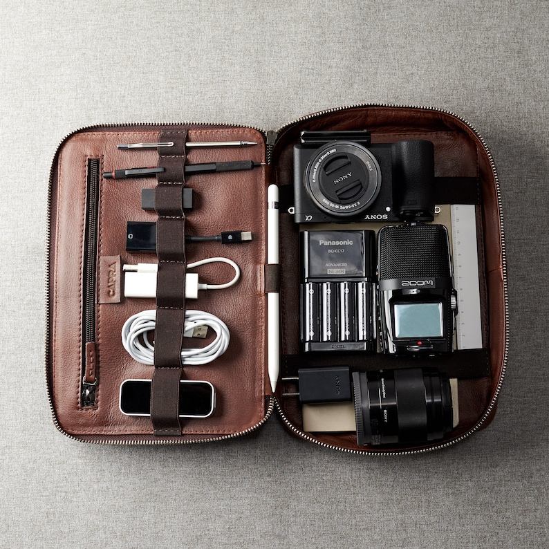 May include: A brown leather carrying case with a zipper closure. The case is open and contains a camera, a microphone, a battery charger, and various cables and accessories.