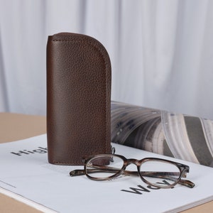 Puede incluir: Un estuche de gafas de cuero marr&oacute;n con cierre de cremallera, junto a unas gafas de pasta marr&oacute;n de carey sobre una revista.