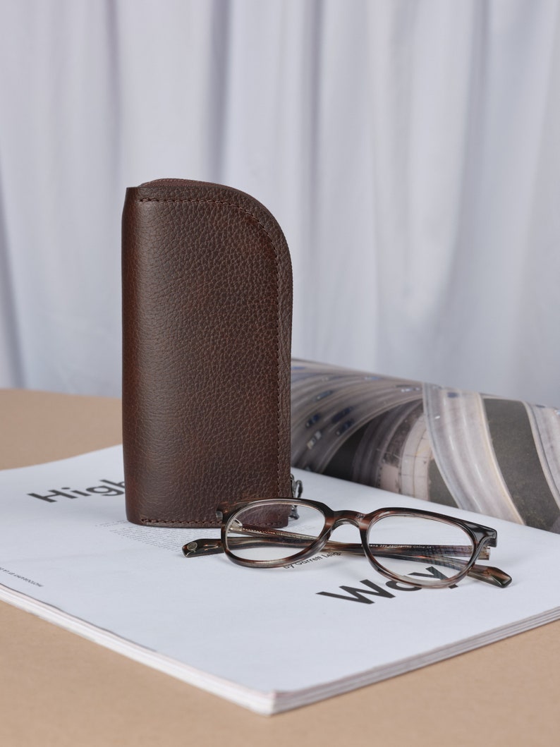 Puede incluir: Un estuche de gafas de cuero marr&oacute;n con cierre de cremallera, junto a unas gafas de pasta marr&oacute;n de carey sobre una revista.