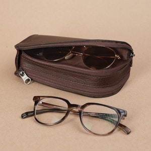 Puede incluir: Un estuche para gafas de cuero marr&oacute;n con cierre de cremallera. El estuche est&aacute; abierto y hay un par de gafas de sol marrones dentro. Un par de gafas de pasta de carey est&aacute;n sobre una superficie marr&oacute;n.