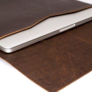 May include: A brown leather laptop sleeve with the word "CAPRA" embossed on the bottom right corner. A silver laptop is partially visible inside the sleeve.