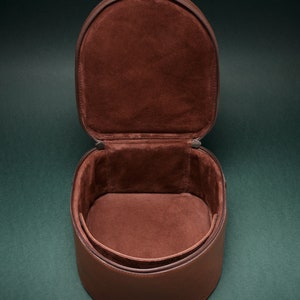 Puede incluir: Una caja redonda de cuero marrón abierta con un interior de ante marrón. La caja es redonda y tiene una cremallera.