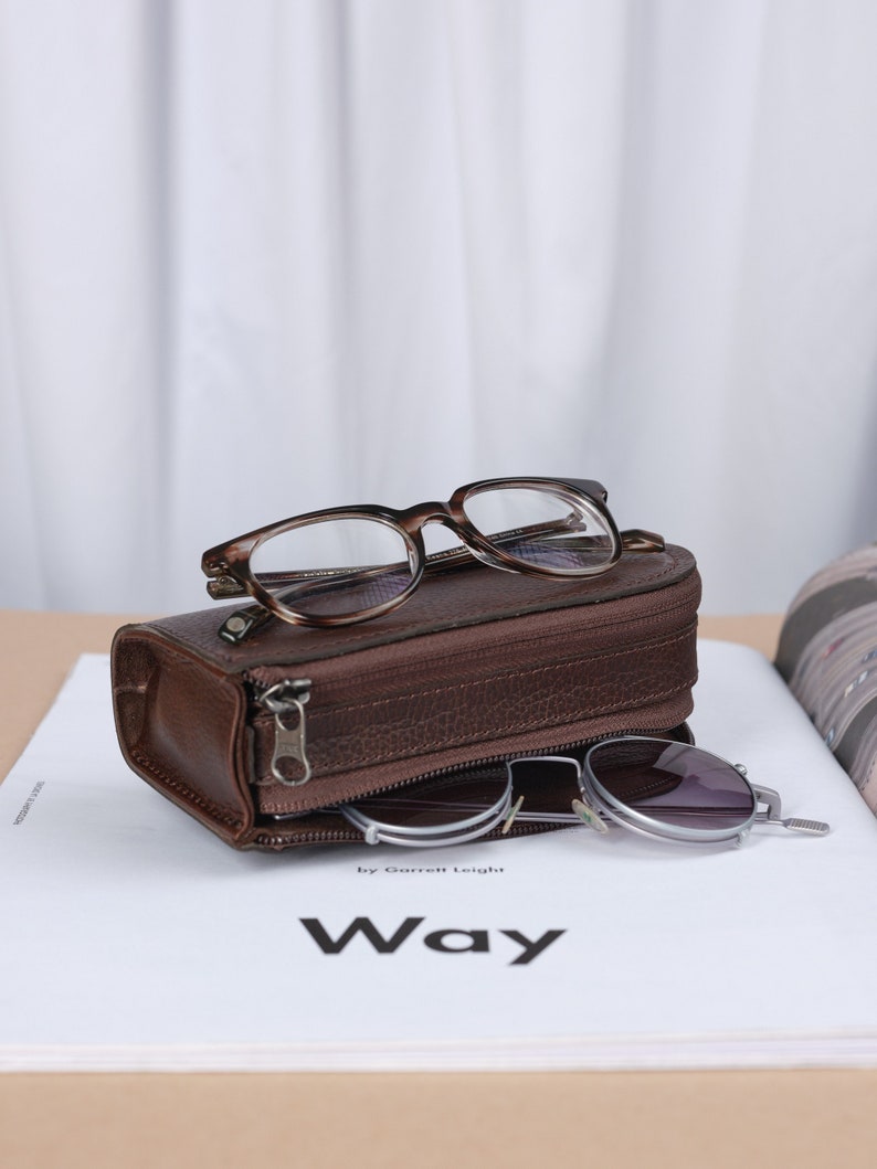 Puede incluir: Un estuche de gafas de cuero marr&oacute;n con cierre de cremallera. El estuche est&aacute; abierto y contiene un par de gafas de sol redondas y un par de gafas rectangulares. El estuche est&aacute; sobre una revista blanca con el texto "Way by Garrett Leigh".