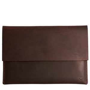 May include: A brown leather laptop sleeve with a flap closure. The sleeve has a subtle distressed look and a small logo on the bottom right corner.