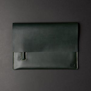 May include: A dark green leather document sleeve featuring a fold-over flap, visible light-colored stitching along the edges, a small integrated pen loop on the front, and a small embossed logo in the bottom right corner of the main body.