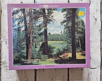 Può includere: Una scatola viola con un puzzle da 1000 pezzi che rappresenta una scena di foresta con un lago e una montagna in lontananza. La scatola riporta la scritta "Guild 1000 Jigsaw Puzzle" e "Mt. Jefferson, Oregon".