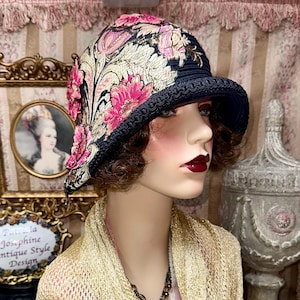 May include: A dark blue cloche hat featuring intricate floral embroidery in shades of pink, cream, and gold. The hat has a structured brim with a decorative braided trim along the edge, showcasing a detailed botanical pattern across the fabric surface.
