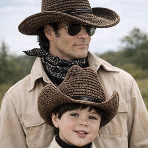 May include: A father and child are wearing matching brown cowboy hats, gloves, and shirts. The hats have a wide brim and a leather band. The child is smiling, and the father is wearing sunglasses. The background is a field.
