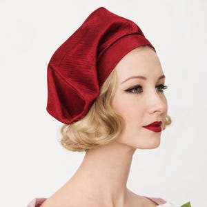 May include: A woman wearing a red, textured hat and a pink dress with a white rose brooch. The hat is a deep red color and has a unique, folded design. The woman has blonde hair and red lipstick.
