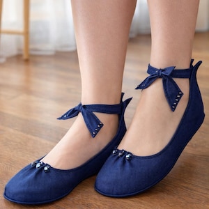 May include: Navy blue ballet flats with ankle straps and bows. The flats have a rounded toe and are embellished with small, silver-colored beads. The ankle straps are adorned with tiny silver studs. The shoes are made of a soft fabric.