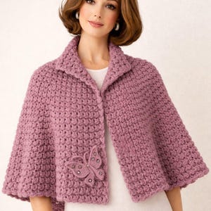 Könnte beinhalten: Ein gehäkelter Umhang in Malvenfarbe mit einer Schmetterlingsapplikation. Der Umhang hat einen Kragen und einen Knopfverschluss. Der Schmetterling ist mit kleinen, glitzernden Akzenten verziert. Das Kleidungsstück ist aus dickem, strukturiertem Garn gefertigt.