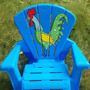 Puede incluir: Una silla de plástico azul para niños con un diseño de gallo. El gallo es verde, amarillo y rojo con lunares blancos.