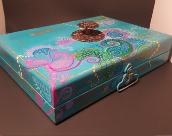 May include: A teal wooden box with a decorative floral design painted on the lid. The box has a metal latch and a metal knob on the top.