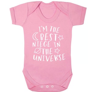 May include: A pink baby bodysuit with the white text "I'M THE BEST NIECE IN THE UNIVERSE". The design includes a moon, star, and planet. It has short sleeves and three poppers at the bottom.
