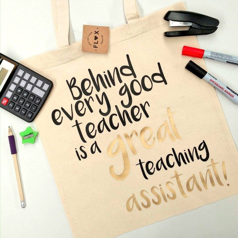 May include: Beige tote bag with the text "Behind every good teacher is a great teaching assistant!" in black and gold lettering. A calculator, stapler, markers, pencil, and sharpener are also visible.