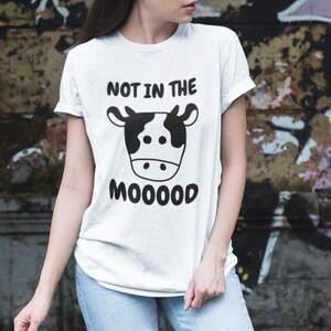 May include: White t-shirt with a black cow graphic and the text "NOT IN THE MOOOOOD". The cow graphic features a black and white cow face. The shirt is made of a soft material.