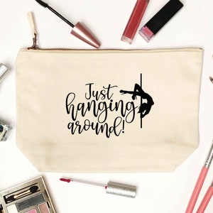 May include: A cream canvas makeup bag with a zip. The bag has the text "Just hanging around!" and a black silhouette of a person on a pole. Lipsticks, brushes, and other makeup items are arranged around the bag on a white surface.