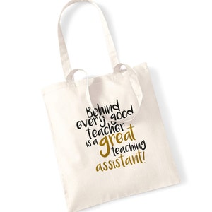 May include: Cream-colored tote bag with the text "Behind every good teacher is a great teaching assistant!" in black and gold lettering. The bag has two long handles and is made of a sturdy fabric, suitable for carrying books or other items.
