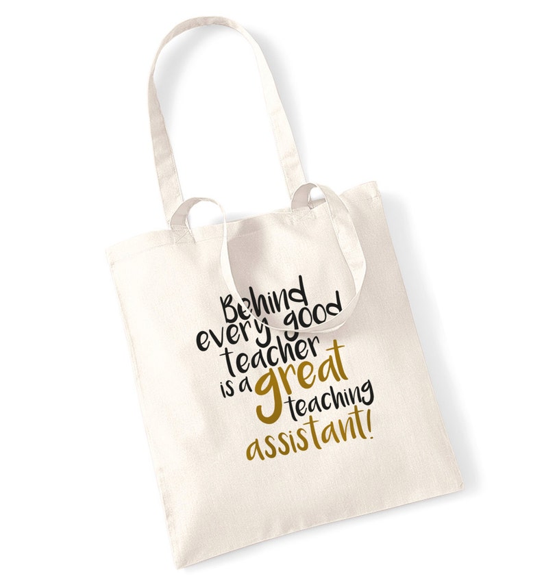 May include: Cream-colored tote bag with the text "Behind every good teacher is a great teaching assistant!" in black and gold lettering. The bag has two long handles and is made of a sturdy fabric, suitable for carrying books or other items.
