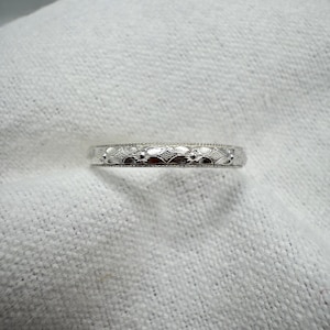 Peut inclure: Une bague en métal argenté présentant un motif gravé répétitif de petites fleurs et de formes en losange le long de l'anneau. Les bords de la bague sont détaillés avec une texture perlée fine, créant un design cohérent sur toute la circonférence.