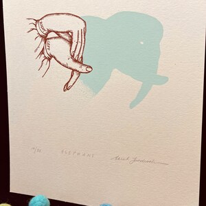 May include: A hand shadow art print with a light blue elephant silhouette. The print is numbered 18/30 and signed "Sarah Janecowski".
