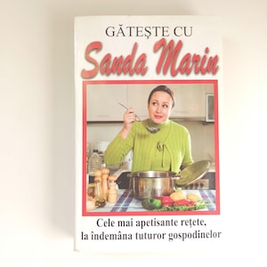 Könnte beinhalten: Ein Kochbuch mit dem Titel "Gătește cu Sanda Marin" zeigt eine Frau in einem grünen Pullover, die in einem Topf rührt. Das Cover hat einen roten Rahmen mit Text auf Rumänisch. Gemüse und Kochutensilien sind sichtbar.