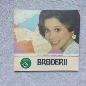 May include: A square book cover titled BRODERII by Liliana Podoleanu and Mihail Popescu. The cover features a smiling person wearing a white collared shirt and a striped earring, set against a background of light blue and pink fabric patterns.