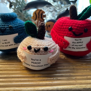 May include: Three crocheted stuffed fruit with Valentine's Day messages. A blue blueberry says "I am so BLUE when I'm not with you." A peach says "You're a PEACH! Happy Valentines Day!" A red apple says "You're the APPLE of my eye! Be My Valentine."
