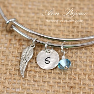 May include: Silver charm bracelet with a silver angel wing charm, a silver charm with the letter "S" and a light blue crystal charm. The bracelet is on a burlap background.