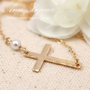 May include: A gold cross necklace with a single white pearl accent. The necklace is on a white fabric background. The text "Ann Bijoux Handcrafted Jewelry Design" is visible at the top of the image.