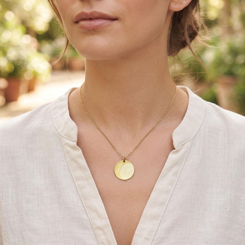 May include: A gold-toned necklace featuring a circular pendant with a textured fingerprint design suspended from a delicate chain. The pendant is round and polished, showcasing a detailed impression on its surface, worn against a white linen shirt.