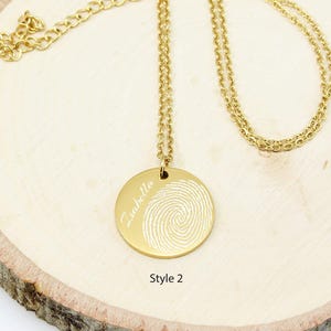 May include: Gold necklace with a round pendant featuring a fingerprint design and the name "Isabella" engraved on it. The pendant is attached to a delicate chain.