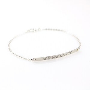 Puede incluir: Una delicada pulsera de plata con un colgante de barra curvada. La barra está grabada con las coordenadas "38° 72'24"W 42° 32'23". La pulsera tiene una fina cadena y un cierre sencillo.