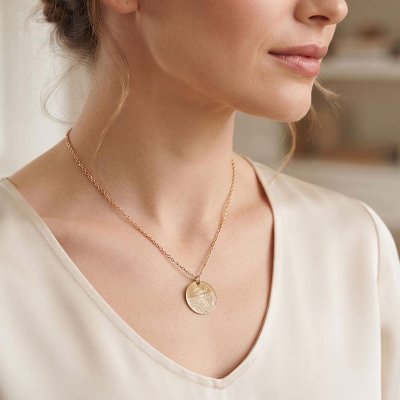 May include: A gold-toned necklace featuring a delicate chain and a circular pendant. The pendant is engraved with a detailed fingerprint pattern and the cursive script name "Dad" positioned horizontally across the center of the circular surface.