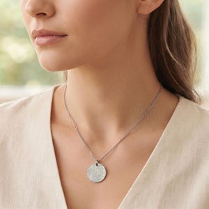 May include: A silver-toned circular pendant necklace featuring a detailed engraved fingerprint pattern on the front surface. The pendant hangs from a delicate matching silver-toned chain that rests against the collarbone area of the wearer.