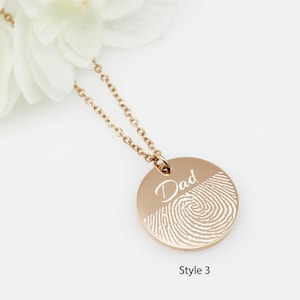 May include: A gold necklace with a round pendant featuring a fingerprint design and the word "Dad" engraved on it. The pendant is on a delicate chain.