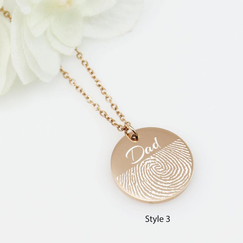 May include: A gold necklace with a round pendant featuring a fingerprint design and the word "Dad" engraved on it. The pendant is on a delicate chain.