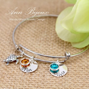 May include: A silver expandable bangle bracelet with three charms. The charms are a small turtle, a round charm with an orange crystal and the name "Erin" engraved on it, and a round charm with a teal crystal and the name "Olivia" engraved on it.