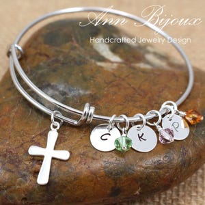 May include: A silver-toned bangle bracelet with a cross charm and several round charms with letters and colorful beads. The bracelet is labeled "Ann Bijoux Handcrafted Jewelry Design."